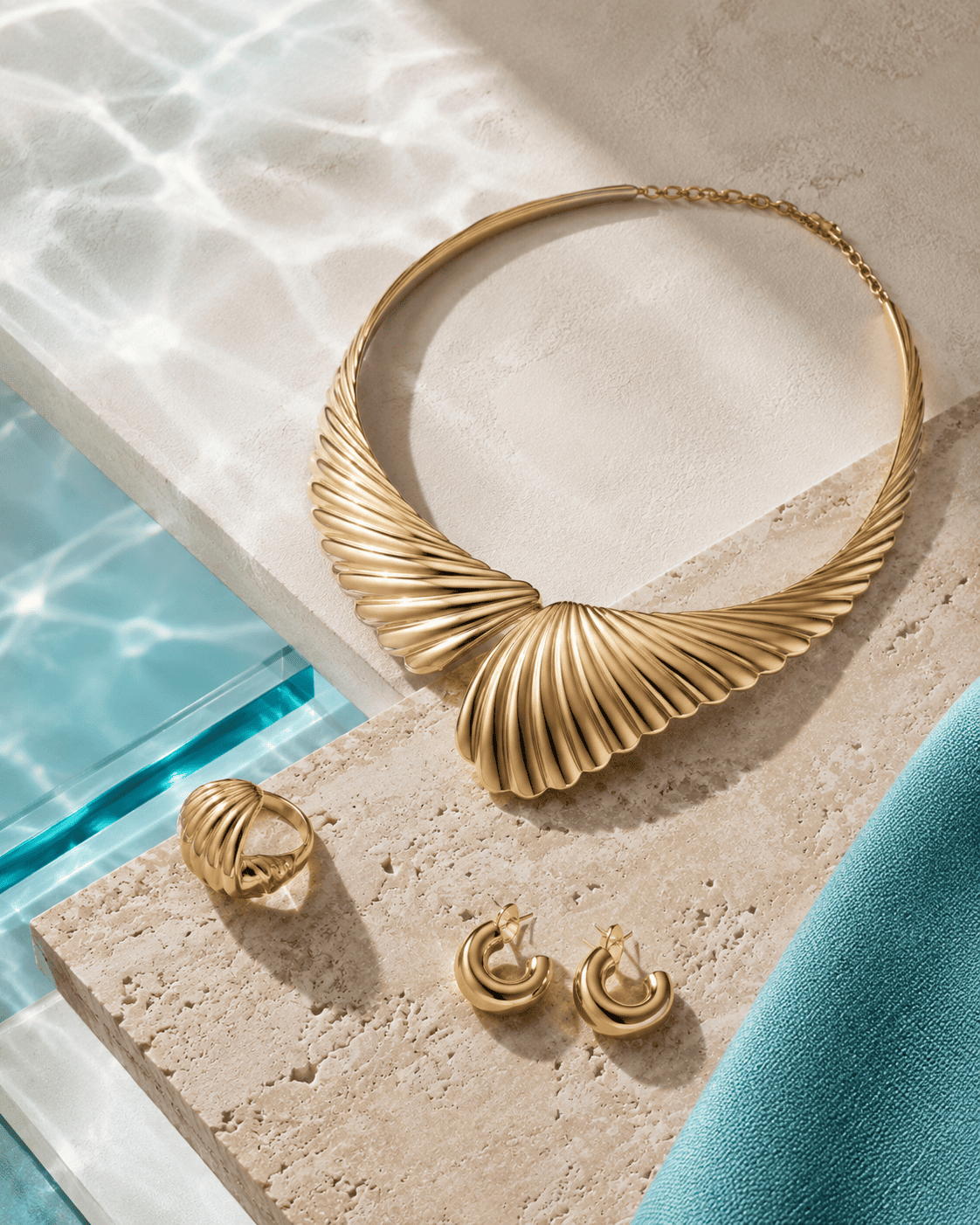 A gold-toned twisted chain necklace with a wide central twist, displayed on a beige stone slab alongside matching gold rings and earrings, with a dark red fabric corner and blue reflective surface in the background.